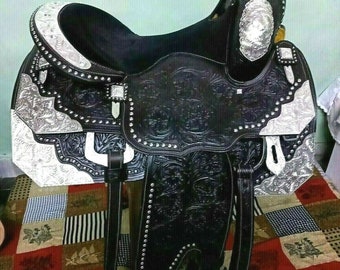May include: A black leather saddle with silver accents and floral tooling. The saddle has a black seat and a silver horn. The saddle is sitting on a patterned rug.