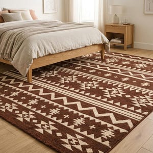 May include: A brown and cream-colored area rug with a geometric pattern is placed in a bedroom. The rug features repeating patterns of zigzags, triangles, and other shapes. A bed with a wooden frame and neutral bedding is in the background.
