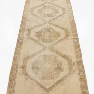 May include: A long, rectangular runner rug featuring a series of repeating geometric diamond and hexagonal motifs in shades of beige, tan, and light brown. The rug has a central pattern of interconnected shapes and a decorative border along the edges.