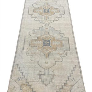 May include: A long, rectangular runner rug featuring a faded beige base with symmetrical geometric patterns in muted blue and tan. The design includes central diamond-shaped medallions with intricate borders and short fringe at both ends.