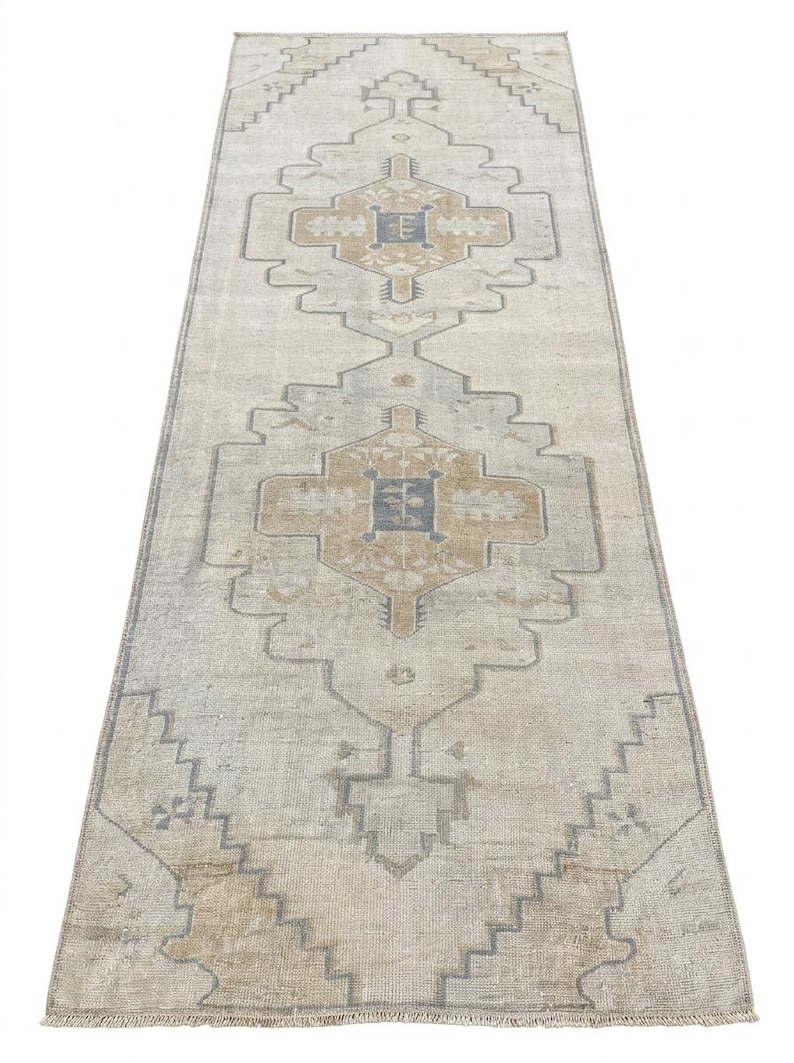 May include: A long, rectangular runner rug featuring a faded beige base with symmetrical geometric patterns in muted blue and tan. The design includes central diamond-shaped medallions with intricate borders and short fringe at both ends.