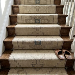 May include: A staircase featuring a long, beige runner rug with a repeating geometric pattern in muted blue and brown tones. The runner is secured to the wooden steps with metallic brass rods. A pair of brown leather slip-on shoes rests on the bottom step.