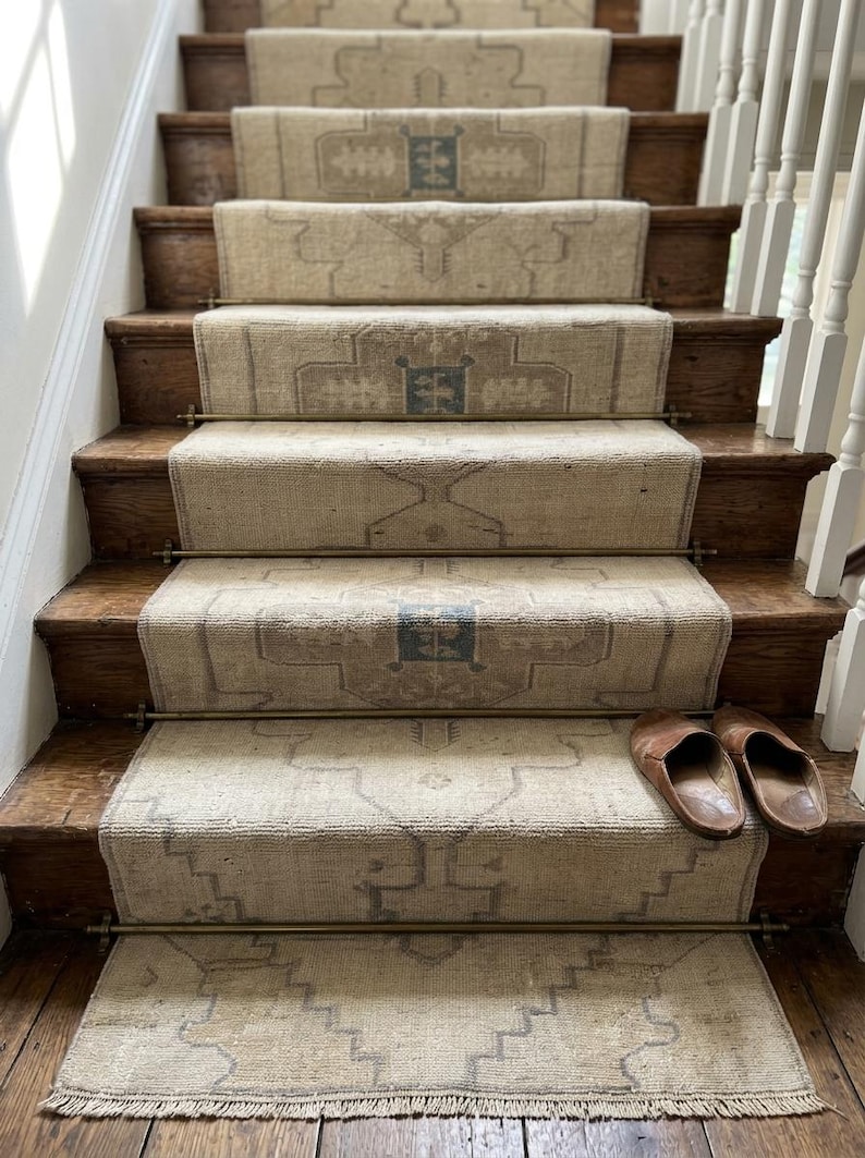 May include: A staircase featuring a long, beige runner rug with a repeating geometric pattern in muted blue and brown tones. The runner is secured to the wooden steps with metallic brass rods. A pair of brown leather slip-on shoes rests on the bottom step.
