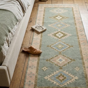May include: A long rectangular runner rug featuring a sage green center field with a repeating pattern of diamonds in shades of blue, tan, and cream. The rug is framed by a wide border in a soft pink hue with small cross motifs and white fringe.