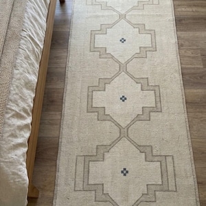 May include: A long, rectangular runner rug in a light beige color featuring a repeating geometric pattern of interconnected diamonds outlined in a darker tan shade, with a small, dark blue cross-shaped motif centered within each diamond shape.
