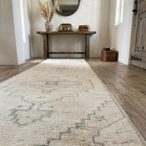 May include: A long, rectangular area rug featuring a cream-colored base with intricate, faded blue geometric patterns and a central medallion design. The rug is positioned on a wooden floor, extending from the foreground toward a wooden console table.