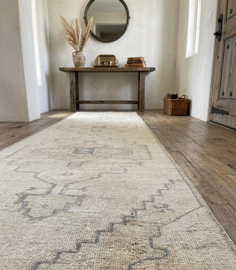 May include: A long, rectangular area rug featuring a cream-colored base with intricate, faded blue geometric patterns and a central medallion design. The rug is positioned on a wooden floor, extending from the foreground toward a wooden console table.
