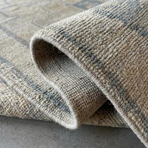 May include: A woven rug featuring a textured, beige and cream pile with a subtle, repeating geometric pattern in muted gray. The rug is folded over to reveal the thick, dense construction of the fibers and the finished, stitched edge detail.
