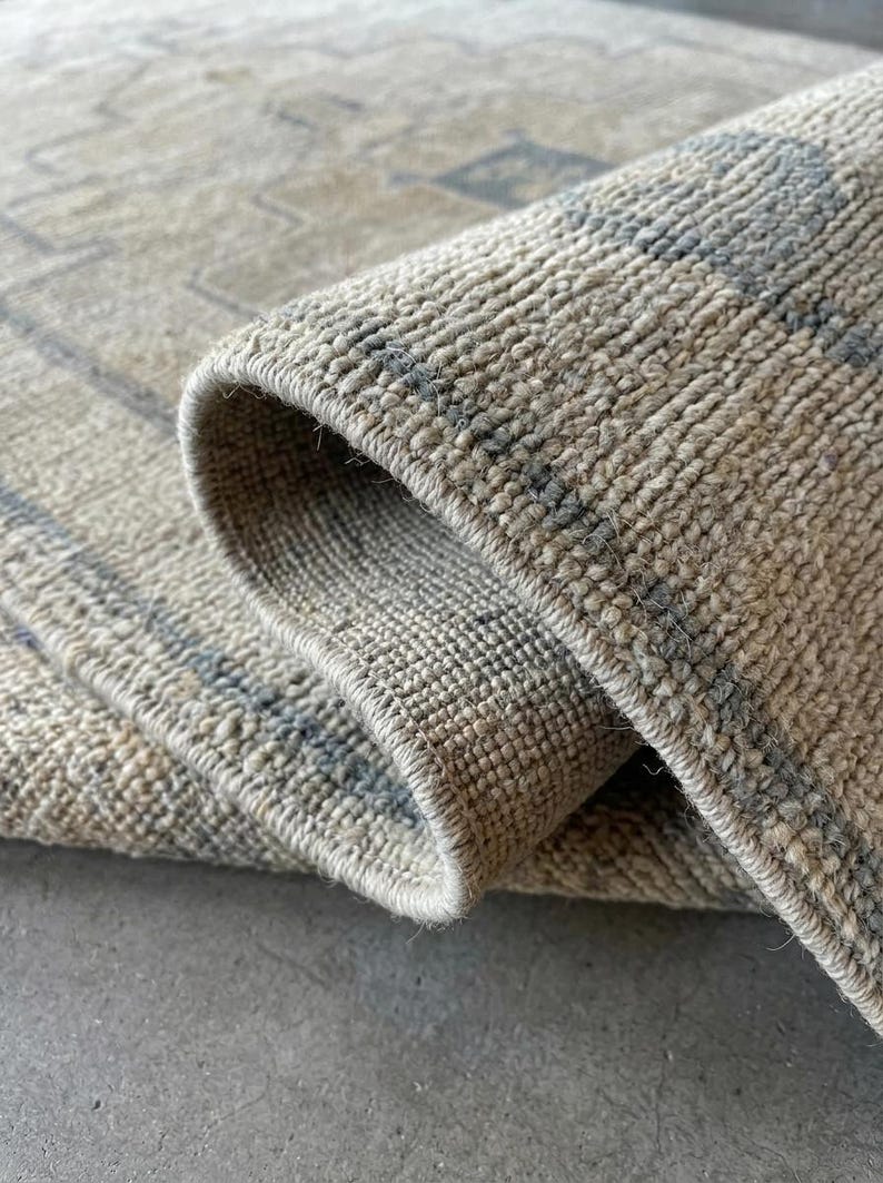 May include: A woven rug featuring a textured, beige and cream pile with a subtle, repeating geometric pattern in muted gray. The rug is folded over to reveal the thick, dense construction of the fibers and the finished, stitched edge detail.