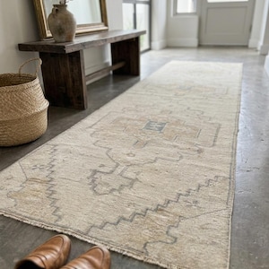May include: A long, rectangular runner rug featuring a faded, neutral-toned pattern with beige, tan, and soft gray geometric motifs. The rug has a low-pile texture, a central medallion design, and short, frayed fringe edges on the ends.
