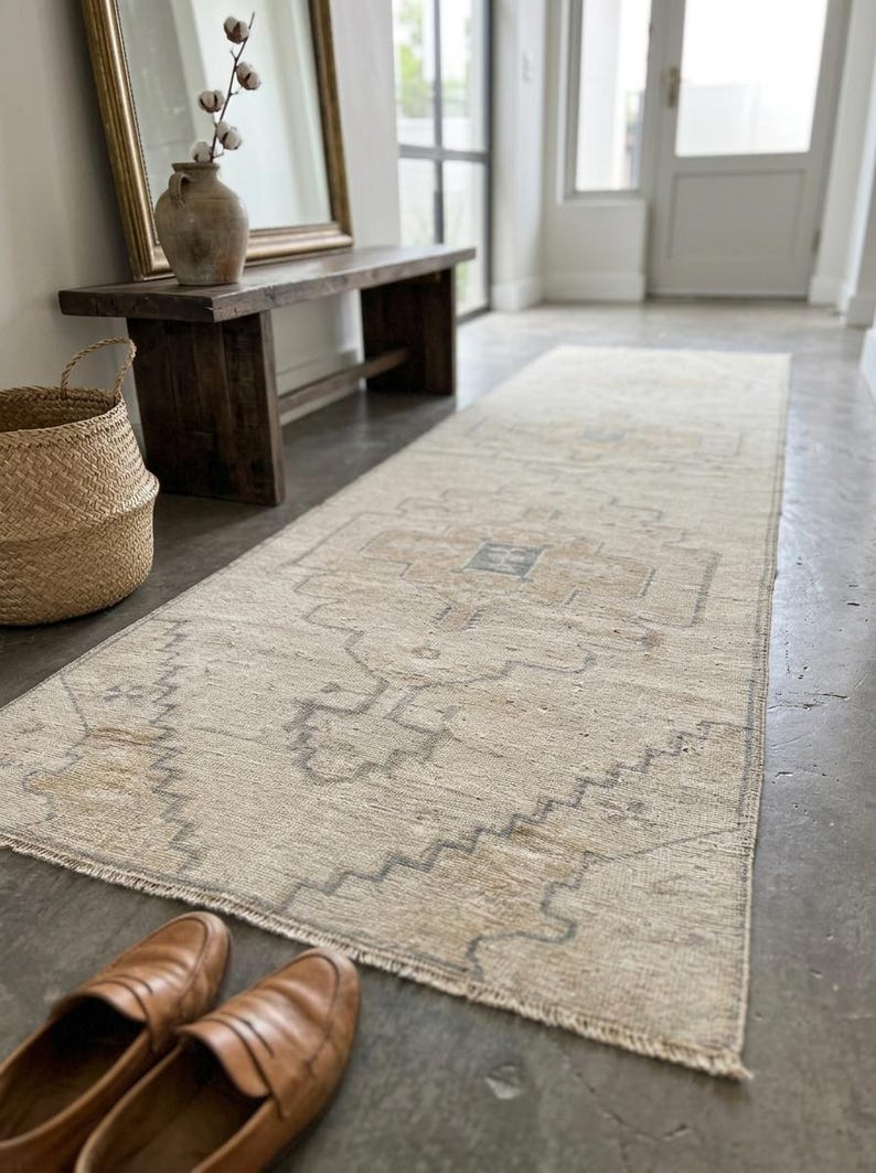 May include: A long, rectangular runner rug featuring a faded, neutral-toned pattern with beige, tan, and soft gray geometric motifs. The rug has a low-pile texture, a central medallion design, and short, frayed fringe edges on the ends.