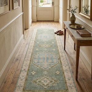 May include: A long, rectangular runner rug featuring a sage green central field with repeating geometric diamond and hexagonal motifs in soft blue, cream, and muted olive tones, finished with a cream-colored border and short fringe at both ends.