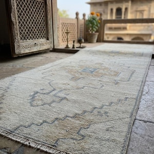 May include: A rectangular runner rug featuring a cream-colored base with a repeating geometric pattern in muted blue and tan tones. The rug has a low-pile texture and short fringe detailing along the edges, showcasing a symmetrical design.