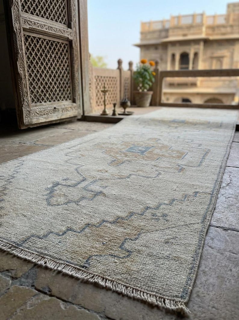 May include: A rectangular runner rug featuring a cream-colored base with a repeating geometric pattern in muted blue and tan tones. The rug has a low-pile texture and short fringe detailing along the edges, showcasing a symmetrical design.