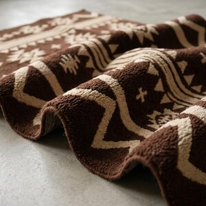 May include: A rolled-up brown and beige patterned rug. The rug features a repeating geometric design in a lighter beige color against a dark brown background. The rug's texture appears soft and plush, suitable for home decor.