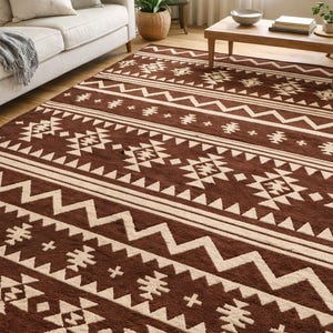 May include: A rectangular brown area rug with a geometric pattern in cream. The design features zigzags, triangles, and stylized shapes arranged in horizontal bands. The rug is placed on a hardwood floor.