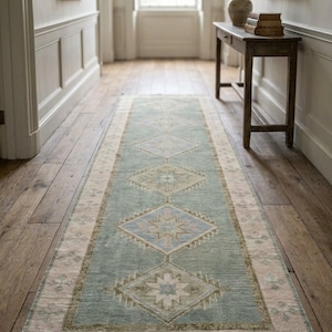 May include: A long, rectangular runner rug featuring a muted sage green central field decorated with a series of repeating diamond-shaped medallions in soft blue, cream, and tan tones, framed by a wide, pale pink border with fringe ends.