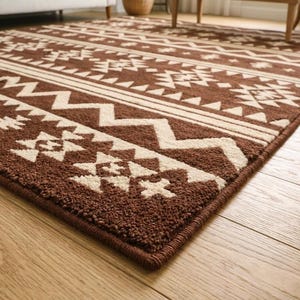 May include: Brown and cream-colored area rug with a geometric pattern. The rug features repeating shapes and lines in a tribal design. The rug is rectangular and has a dark brown border.