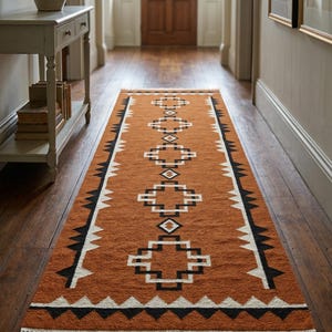 May include: An orange and white geometric patterned runner rug with a fringed edge. The rug features a repeating pattern of diamonds and crosses in white and black. The rug is placed on a wooden floor.
