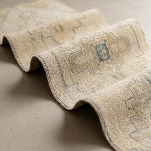 May include: A beige runner rug featuring a repeating geometric pattern in muted blue and yellow tones. The rug is folded in a wave-like shape, showcasing its textured surface and the intricate, symmetrical design elements woven into the fabric.