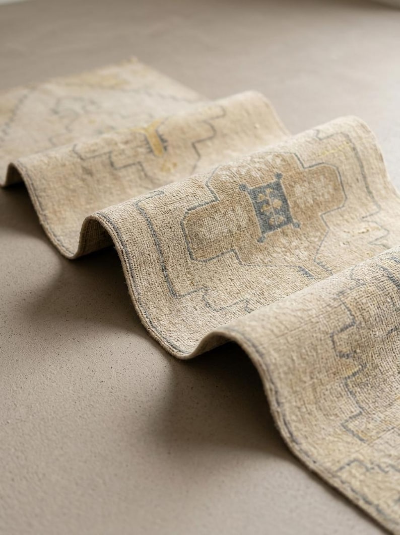 May include: A beige runner rug featuring a repeating geometric pattern in muted blue and yellow tones. The rug is folded in a wave-like shape, showcasing its textured surface and the intricate, symmetrical design elements woven into the fabric.