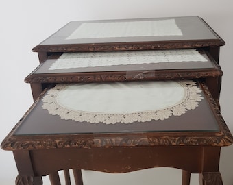 May include: Set of three nesting tables with dark brown wooden frames and glass tops. Each table has a decorative, carved edge and a white lace doily. The tables are nested together, with the smallest table at the bottom.