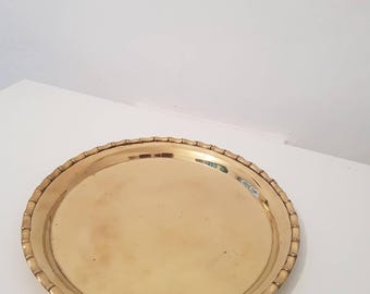 May include: A round, gold-colored metal tray with a decorative, ridged edge. The tray is empty and sits on a white surface. The tray's surface reflects light, and the overall aesthetic is vintage or antique.