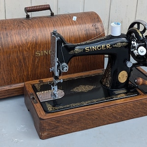 May include: A vintage black Singer sewing machine with a wooden case. The machine has a gold-coloured design on the front and a spool of white thread on top.