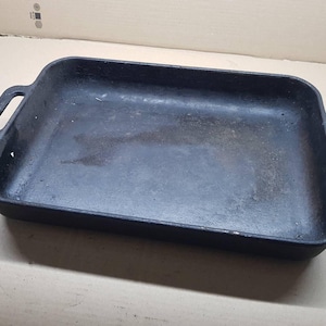 May include: A rectangular, black cast iron baking tray with two handles. The tray has a slightly worn appearance, suggesting it has been used. The handles are rectangular and integrated into the tray's design. Suitable for baking and roasting.