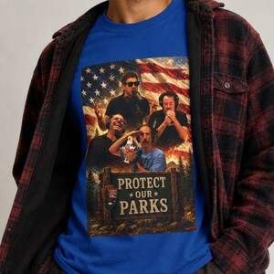 May include: A brown knit beanie, a blue t-shirt with a graphic print of four people, an American flag, and the text "PROTECT OUR PARKS", and a red and black plaid long-sleeve button-down shirt worn over the t-shirt with dark grey trousers.