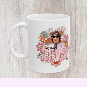 May include: White ceramic mug with a retro-style design featuring a man wearing sunglasses and headphones, a microphone, and flowers. The text "PRAISE GOD BABY" is printed in pink.