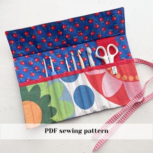 Op de afbeelding: Een kleurrijke haaknaaldrol met een blauwe appelprint bovenkant, rode rand en een patroon aan de voorkant met groene, blauwe en rode cirkels. De rol bevat haaknaalden en een witte schaar. De tekst "PDF sewing pattern" is zichtbaar.