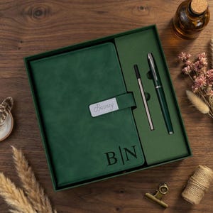 May include: A dark green journal and pen set in a gift box. The journal has the name "Barney" on a silver plate and initials "B|N" embossed on the cover. A silver pen and refill are included. The set is displayed on a wooden surface.