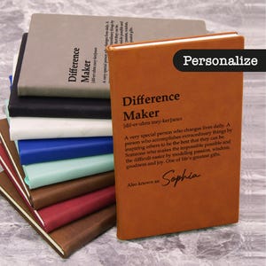 May include: A stack of leather-bound journals in various colors, including brown, black, and blue. The front journal is a warm brown with the word "Difference Maker" and the name "Sophia" printed on it. The word "Personalize" is also visible.