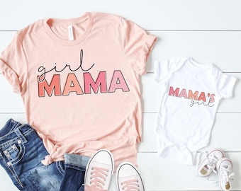 May include: Pink and white t-shirts with the text "girl mama" and "mama's girl" in a gradient pink and orange font. The shirts are on a white wooden background with a pair of pink shoelace sneakers.