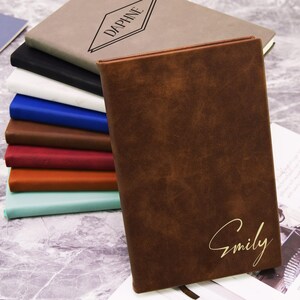 May include: A stack of personalized notebooks in various colors, including brown, black, white, blue, and red. The front notebook is brown with the name "Emily" in gold script. The top notebook is tan with the name "Daphne" in a black diamond.