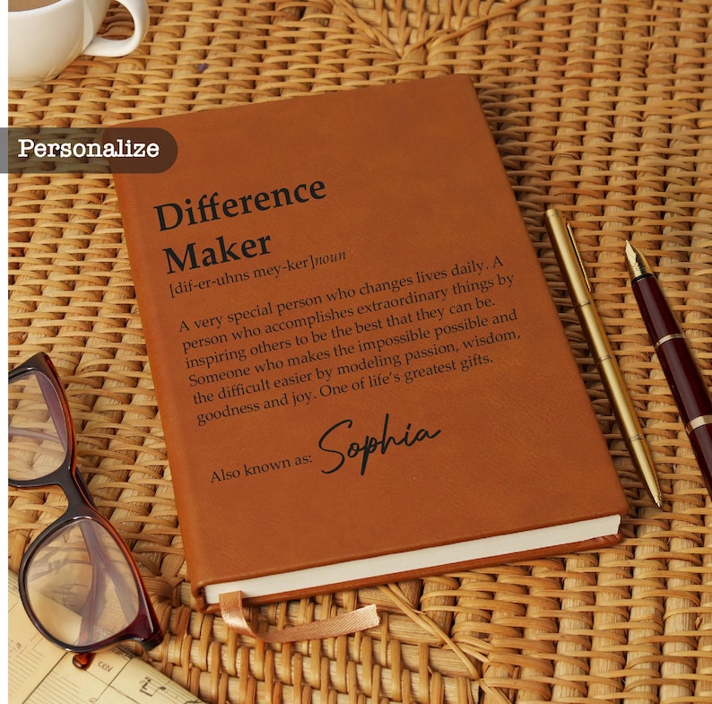 May include: A brown leather journal with the words "Difference Maker" and a definition printed on the cover. The journal is personalized with the name "Sophia". A gold pen, a burgundy pen, and glasses are next to the journal.