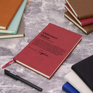 May include: A red journal with the text "Difference Maker" and a definition. The journal has a red ribbon bookmark and the name "Sophia" printed on it. Other journals in various colors are in the background.
