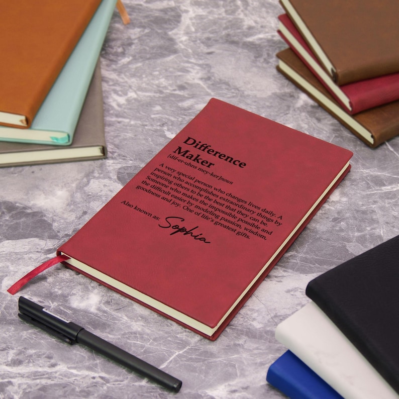 May include: A red journal with the text "Difference Maker" and a definition. The journal has a red ribbon bookmark and the name "Sophia" printed on it. Other journals in various colors are in the background.