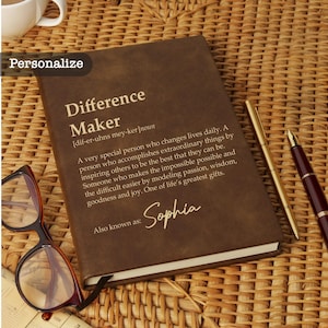 May include: A brown leather-bound journal with the words "Difference Maker" and a definition printed on the cover. The journal includes the name "Sophia" and is next to a pair of glasses and pens.