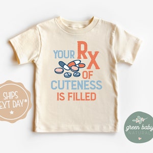 Puede incluir: Camiseta crema para ni&ntilde;os peque&ntilde;os con el texto "YOUR RX OF CUTENESS IS FILLED" en naranja y azul, con gr&aacute;ficos de pastillas. La camiseta de manga corta tiene cuello redondo.