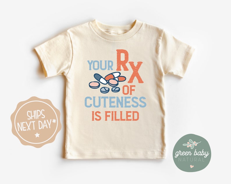 Puede incluir: Camiseta crema para ni&ntilde;os peque&ntilde;os con el texto "YOUR RX OF CUTENESS IS FILLED" en naranja y azul, con gr&aacute;ficos de pastillas. La camiseta de manga corta tiene cuello redondo.