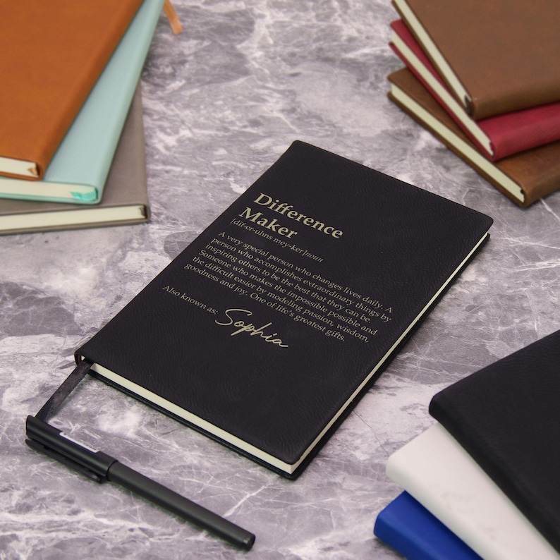 May include: Black notebook with the text "Difference Maker" and a definition. The notebook also has the name "Sophia" printed on it. A black pen rests beside the notebook. Other notebooks are visible in the background.