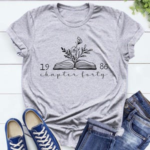 May include: Athletic heather gray t-shirt with a black floral and book design. The text on the shirt reads "1986 chapter forty". Blue canvas sneakers and denim jeans are also visible.
