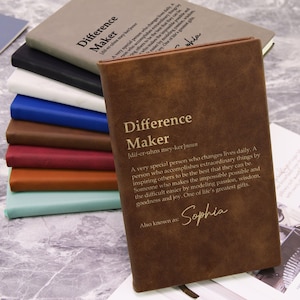 May include: A stack of notebooks in various colors, including brown, black, white, blue, and teal. The front notebook is brown with the words "Difference Maker" and a definition printed on the cover. The name "Sophia" is also printed on the cover.