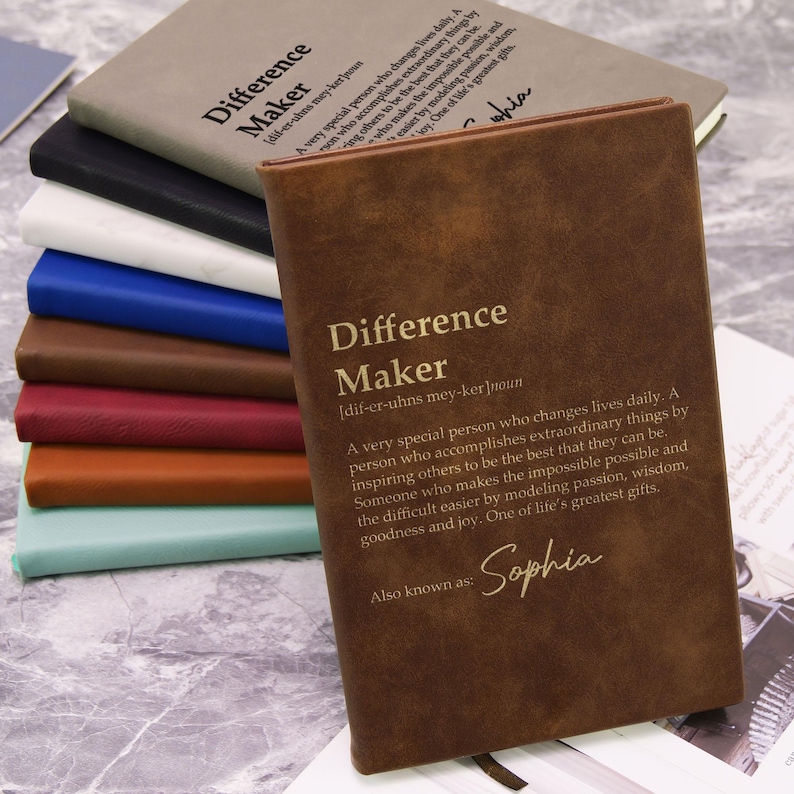 May include: A stack of notebooks in various colors, including brown, black, white, blue, and teal. The front notebook is brown with the words "Difference Maker" and a definition printed on the cover. The name "Sophia" is also printed on the cover.