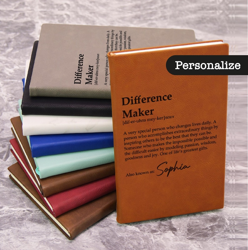 May include: A stack of leather-bound journals in various colors, including brown, black, and blue. The front journal is a warm brown with the word "Difference Maker" and the name "Sophia" printed on it. The word "Personalize" is also visible.