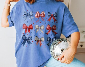 May include: A blue t-shirt featuring a pattern of patriotic bows in red, white, and blue. The bows are decorated with stars and stripes. The shirt is a comfortable, casual style.