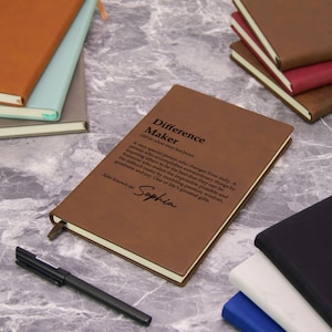 May include: A brown leather journal with the text "Difference Maker" and a definition. The journal has a ribbon bookmark and is next to a black pen. Other journals in various colors are in the background.