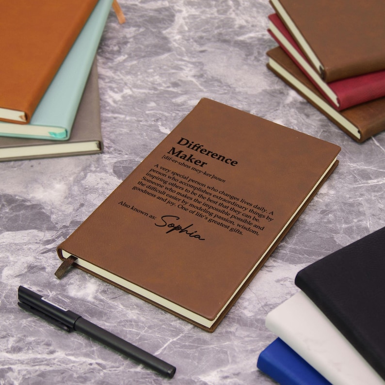 May include: A brown leather journal with the text "Difference Maker" and a definition. The journal has a ribbon bookmark and is next to a black pen. Other journals in various colors are in the background.
